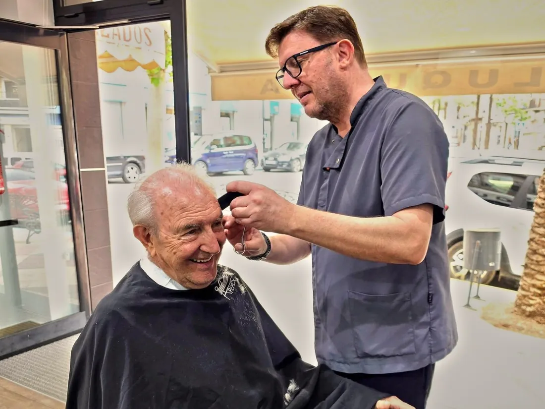 El barbero de Rapiño corta con tijera y peine a un cliente mayor que sonríe en el sillón de la peluquería, en Aspe.
